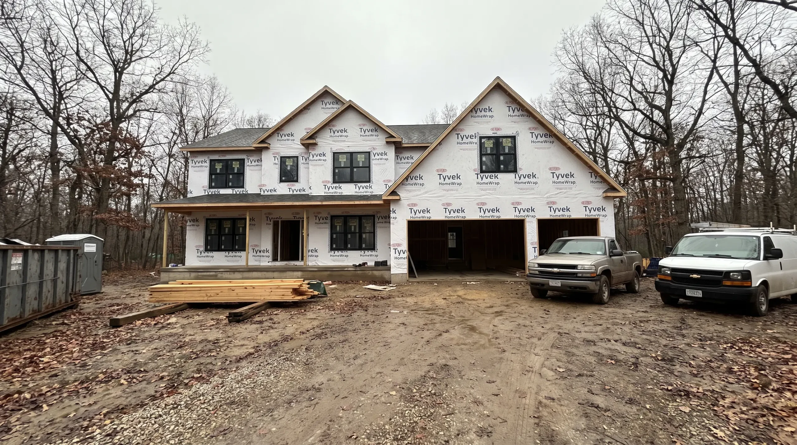 Large new home under construction on a wooded lot, in house wrap, with trucks and a dumpster on the drive