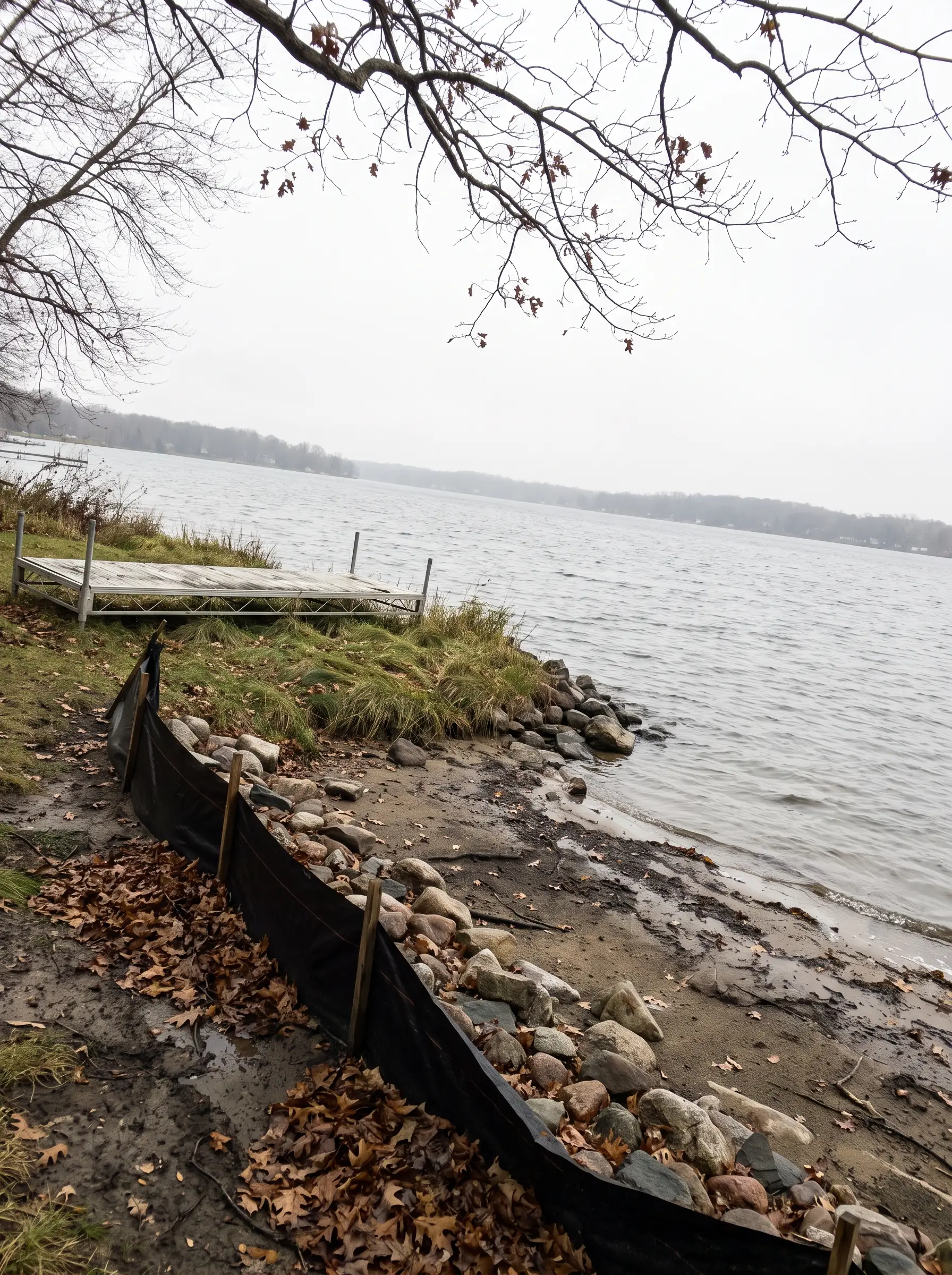 A stony lake shoreline with a black silt fence staked along the bank and a dock section pulled up on the grass