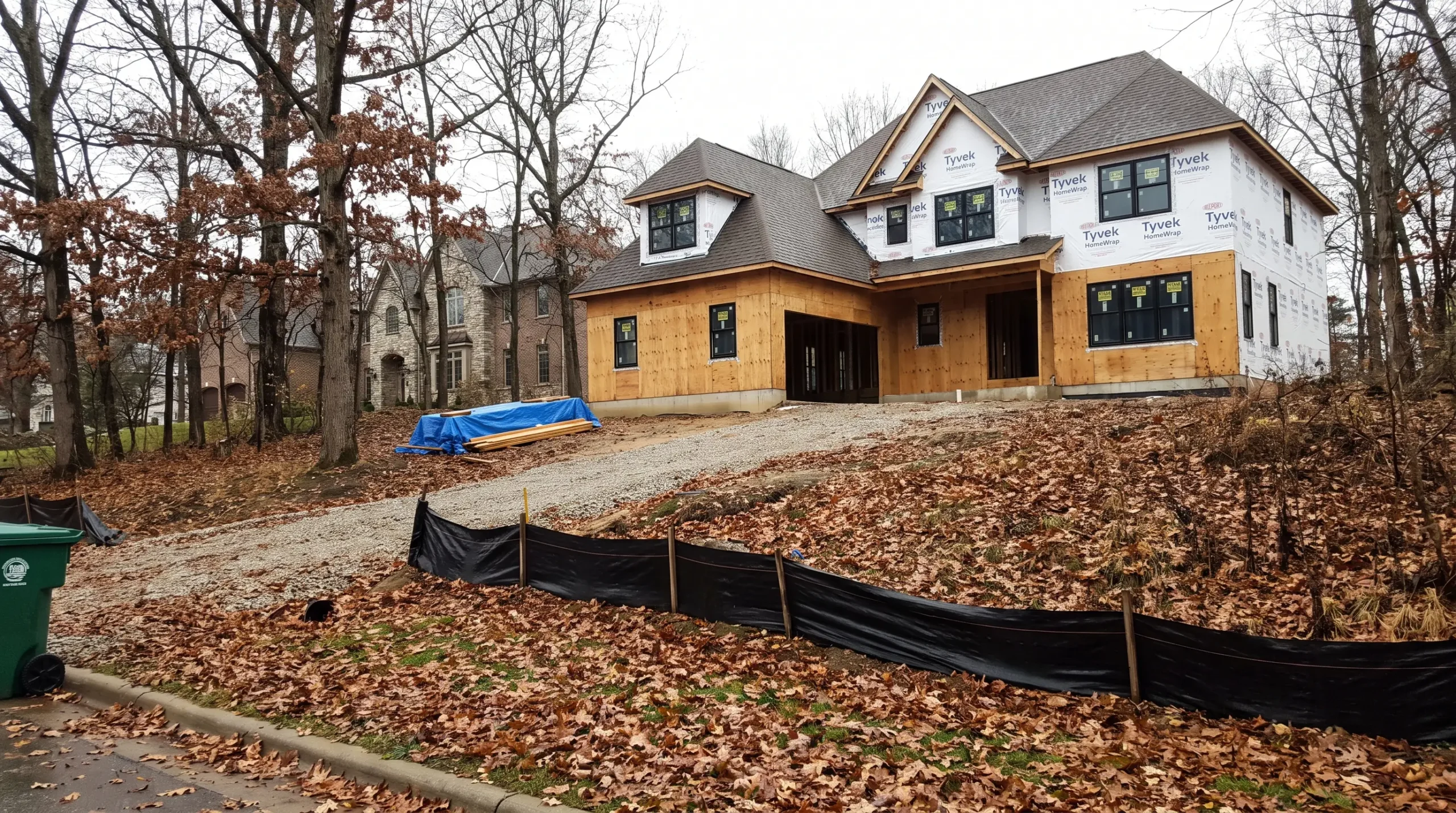 Large custom home under construction on a wooded hillside lot with a silt fence across the slope