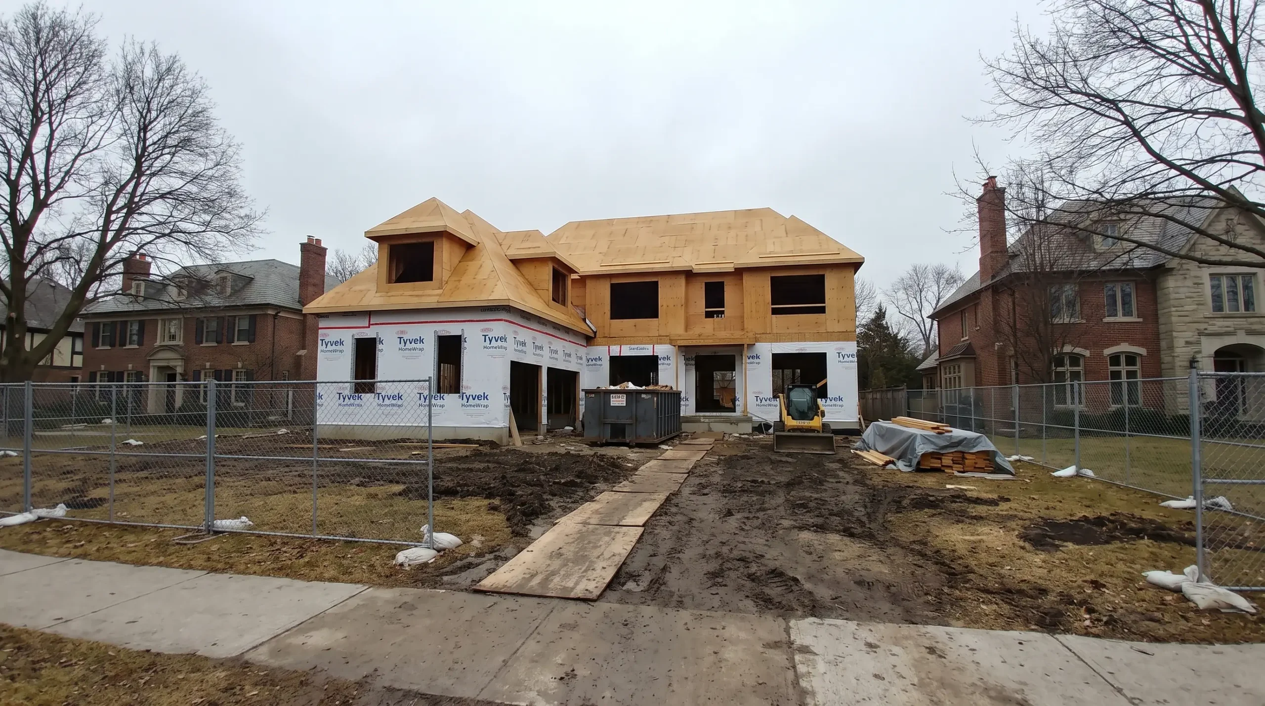A large new home under construction set far back behind a muddy front yard, between older brick and stone estate homes