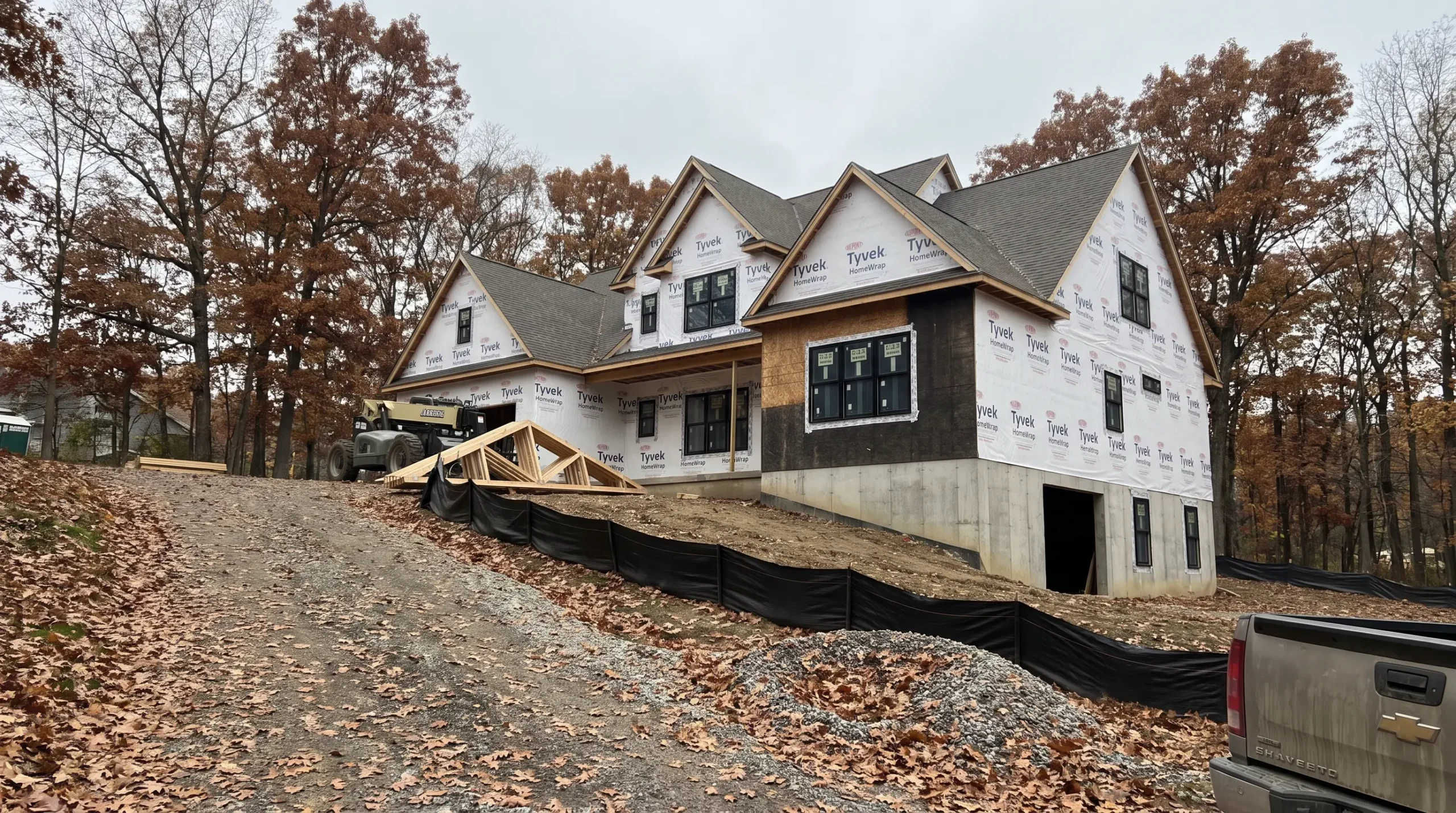 A large custom home under construction at the top of a gravel driveway on a wooded hillside, with roof trusses stacked beside it