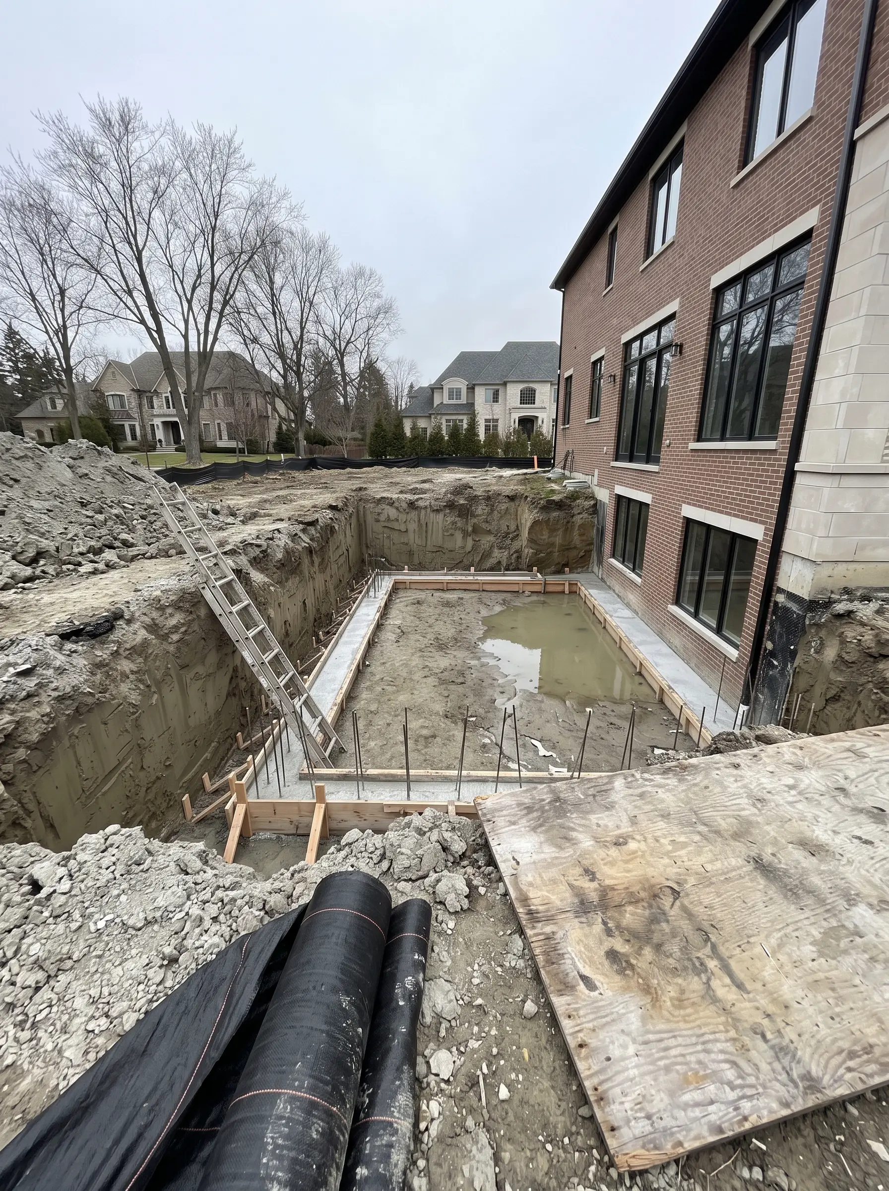 A freshly dug basement excavation with new concrete footings and a ladder, beside the brick wall of a large neighboring home
