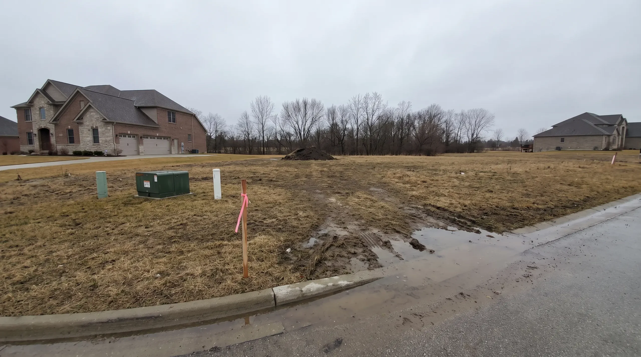 An empty staked home lot with a utility box at the curb, a test hole mound and large finished homes on the neighboring lots